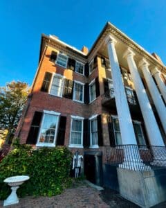 An angled side view of the Andrew-Safford House in Salem Massachusetts, highlighting the white columns, red brick walls, black shutters, and vibrant ivy near a white garden urn.