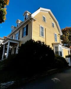 Angled side view of the John P. Peabody House in Salem Massachusetts, featuring its yellow siding, white trim, dormer windows, and leafy surroundings.