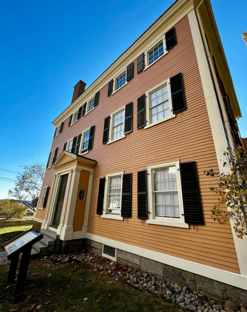 Angled view of the Hawkes House in Salem, Massachusetts, showcasing its peach clapboard siding, black shutters, white trim, and a historical marker in front of the building under a clear blue sky.