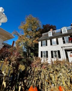 The Ropes Mansion in Salem, Massachusetts, decorated for autumn with corn stalks and pumpkins in front of a white colonial house surrounded by trees with fall foliage under a clear blue sky.