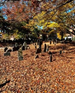 Gravestones at the Old Burying Point cemetery in Salem Massachusetts surrounded by vibrant autumn trees and a thick carpet of fallen leaves in warm orange and brown tones.