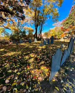 Fallen autumn leaves blanket the grass among gravestones in Old Burying Point Cemetery, Salem Massachusetts, surrounded by trees with colorful foliage under a bright blue sky.
