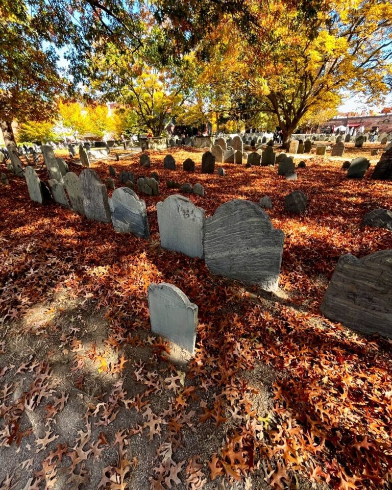 Weathered gravestones surrounded by a vibrant carpet of orange and red autumn leaves at Old Burying Point Cemetery in Salem Massachusetts, with trees in golden foliage creating a peaceful atmosphere.