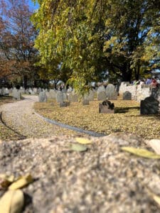 The Old Burying Point Cemetery in Salem, Massachusetts, with gravestones among fallen autumn leaves, a winding path, and sunlight filtering through the trees.