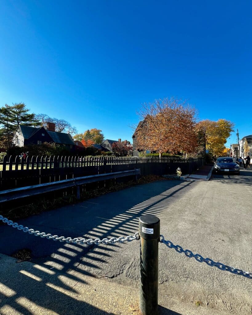 A bright autumn day near the House of the Seven Gables in Salem, Massachusetts, featuring a chain-link barrier, a black wooden fence, and colorful trees under a clear blue sky.