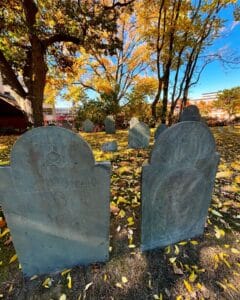 Two gravestones in Salem Massachusetts Charter Street Cemetery, surrounded by yellow and orange autumn leaves, with tall trees in the background under a bright blue sky.