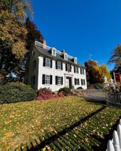 The Ropes Mansion in Salem, Massachusetts, a white colonial house with black shutters, surrounded by vibrant autumn foliage, a white picket fence, and a lawn covered in golden leaves under a clear blue sky.