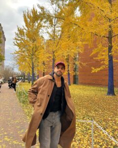 A man in a brown coat and red cap stands on a path covered in yellow leaves at the Peabody Essex Museum in Salem, Massachusetts. The trees around him have golden foliage and painted blue trunks, with a brick building in the background.