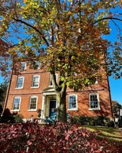 The Merchant Hotel in Salem, Massachusetts, framed by a large tree with bright autumn leaves and surrounded by red bushes and a green lawn.