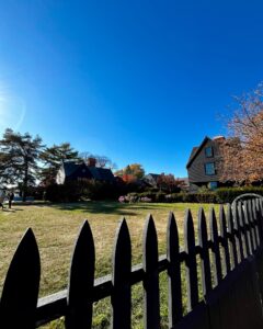 A picturesque garden scene at the House of the Seven Gables in Salem, Massachusetts, featuring a black picket fence, grassy lawn, and vibrant autumn foliage under a bright blue sky.