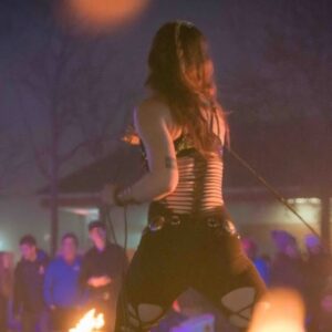 A fire performer at the Frozen Fire Festival in Salem, Massachusetts, seen from behind, wearing a dramatic costume and preparing for her captivating fire act under the evening sky.