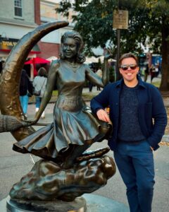 A man smiling and posing beside the bronze "Bewitched" statue of actress Elizabeth Montgomery, who played Samantha in the popular television show, in Salem, Massachusetts.