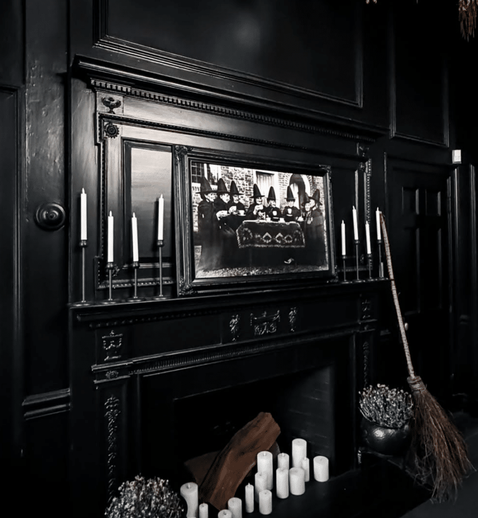 Ornate black fireplace in Salem, Massachusetts, decorated with tall white candles, a vintage broomstick, and a haunting photograph of witches gathered around a table.