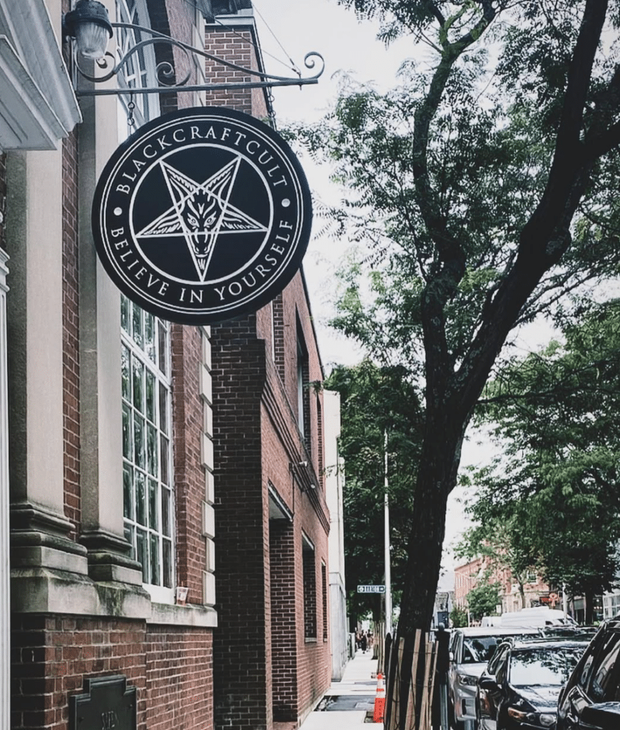 Blackcraft Cult sign featuring a pentagram and the words "Believe in Yourself," displayed on a historic brick building along a tree-lined street in Salem, Massachusetts.