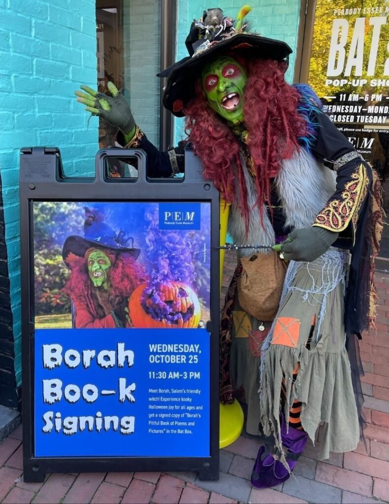 Borah, the vibrant green-faced witch with flowing red hair, stands beside a sign advertising her book signing at the Peabody Essex Museum in Salem, Massachusetts.