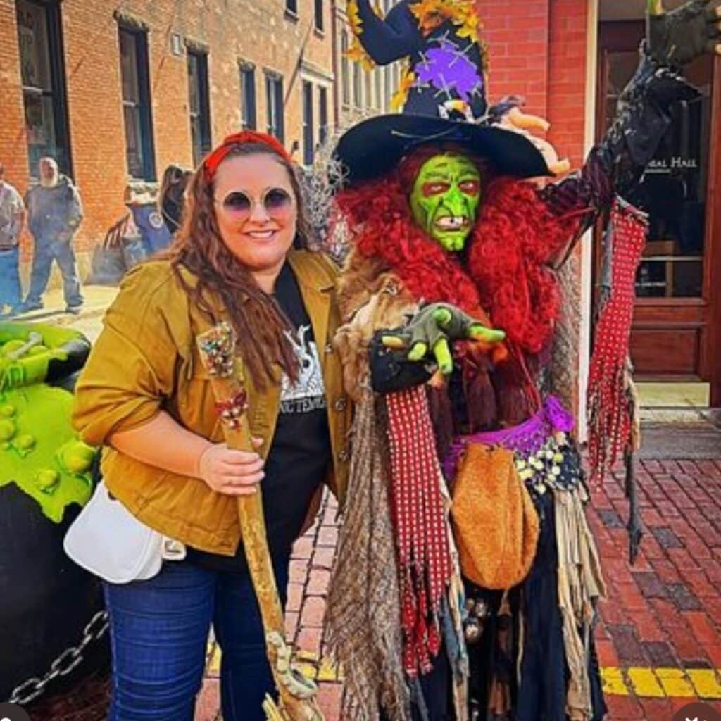 Borah, dressed as a vibrant green-faced witch with red hair, poses with a Halloween enthusiast holding a staff in Salem, Massachusetts.