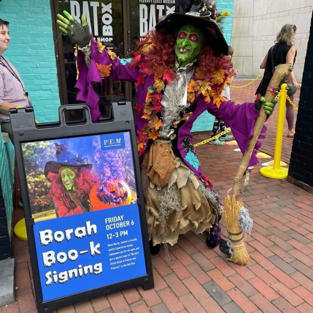 Borah, the green-faced witch with vibrant red hair, poses beside a sign advertising her book signing event outside the Peabody Essex Museum in Salem, Massachusetts.