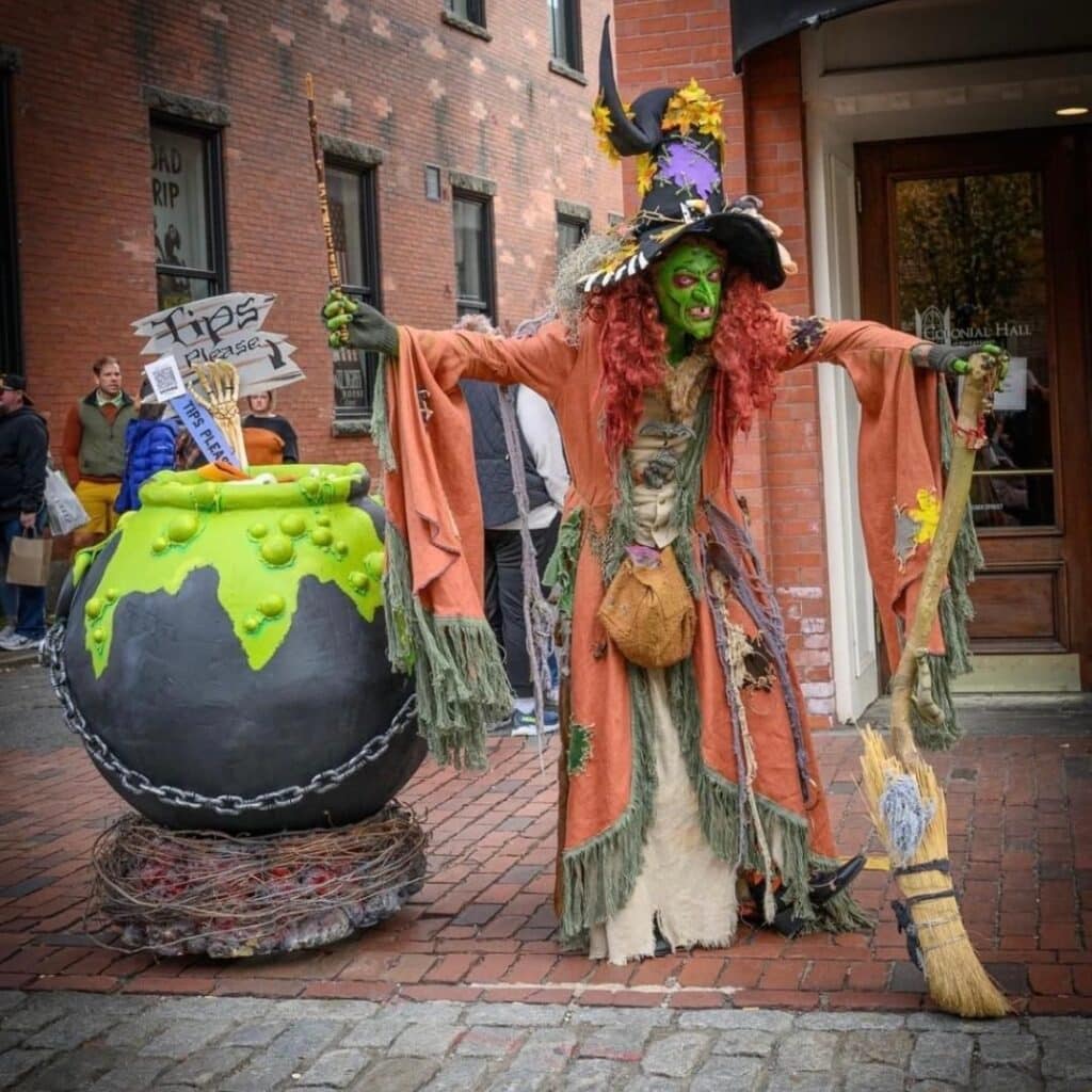Borah, a green-faced witch in an orange tattered cloak, stands with her broom beside a large, bubbling black cauldron decorated with neon green slime and chains in Salem, Massachusetts.