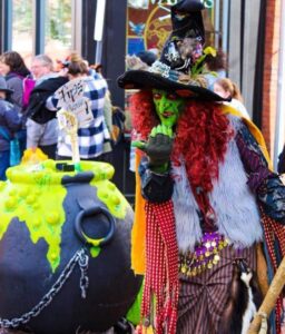 Borah, a vibrant green-faced witch with flowing red hair, stands beside her bubbling black cauldron covered in neon green "slime" and chains, entertaining a crowd in Salem, Massachusetts.