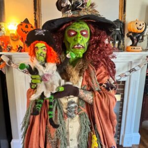 Borah, a witch in Salem, Massachusetts, with vibrant green face paint and red hair, holds a matching miniature doll in a festive Halloween setting.