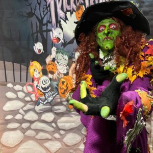 Borah, a witch with green face paint and red hair, poses in front of a colorful Halloween backdrop featuring cartoon characters with pumpkins in Salem, Massachusetts.