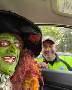 Borah, a witch with green face paint and red hair, grins from a car window while a friendly Salem police officer leans in, sharing a lighthearted moment in Salem, Massachusetts.