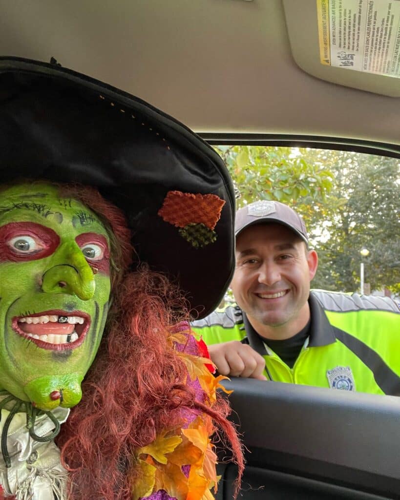 Borah, a witch with green face paint and red hair, grins from a car window while a friendly Salem police officer leans in, sharing a lighthearted moment in Salem, Massachusetts.