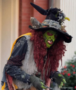 Borah, a witch in Salem, Massachusetts, with green face paint, red hair, and an intricate hat, leans forward with a sly expression during a festive event.