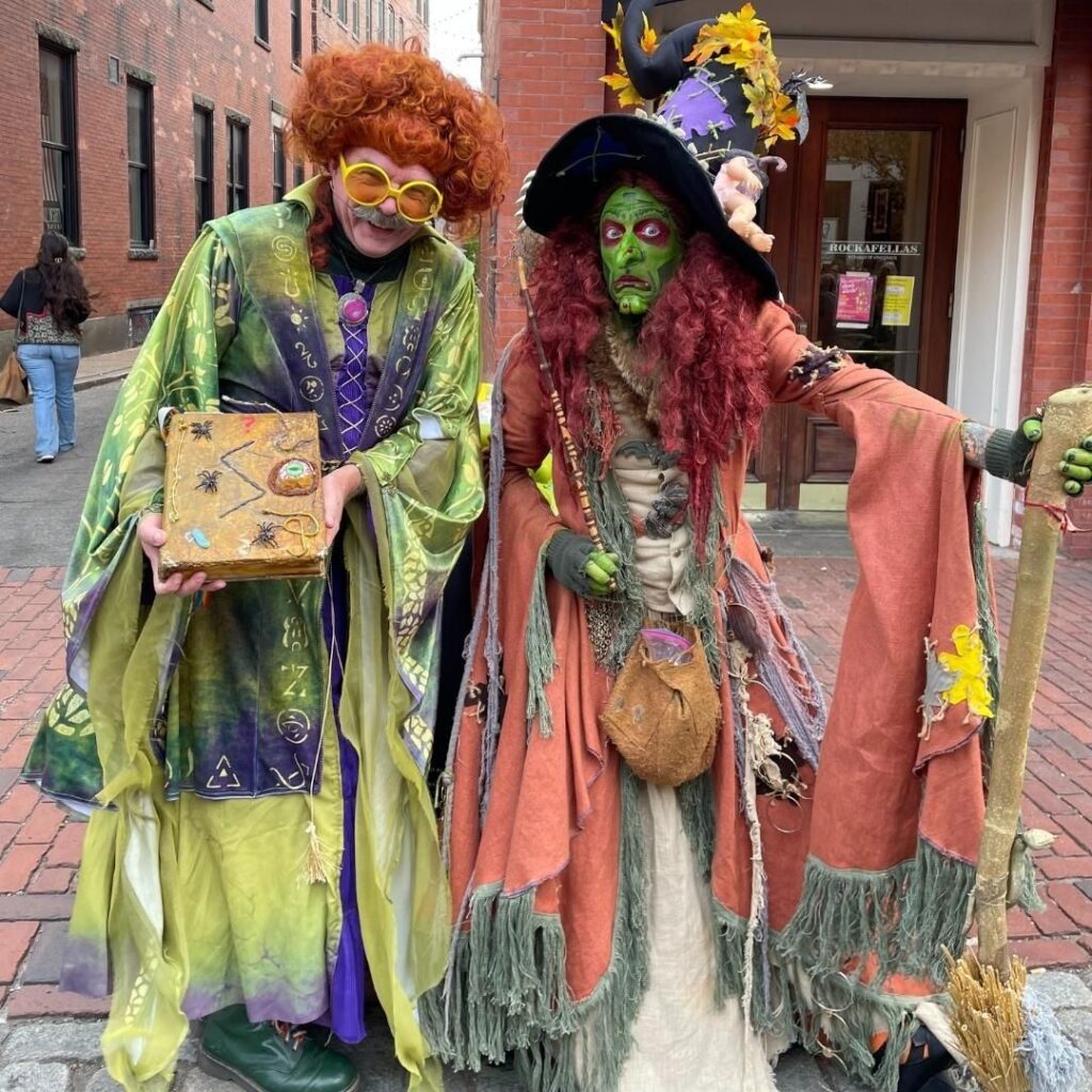 Borah, a witch with green face paint and red hair, stands alongside Winifred Sanderson, a character dressed in vibrant green robes with red curly hair, on the cobblestone streets of Salem, Massachusetts.
