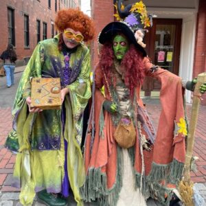 Borah, a witch with green face paint and red hair, stands alongside Winifred Sanderson, a character dressed in vibrant green robes with red curly hair, on the cobblestone streets of Salem, Massachusetts.