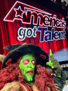 Borah, a witch with green face paint and red hair, poses excitedly in front of the America's Got Talent sign, showcasing her unique style in Salem, Massachusetts.