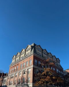 A large brick building with dormer windows stands at the corner of Washington and Essex Street in Salem, Massachusetts. The bright blue sky contrasts with the building's red brick exterior and green roof.