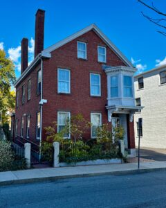 A historic red brick house in Salem, Massachusetts, featuring a white bay window, three chimneys, and a small front garden enclosed by a black iron fence.
