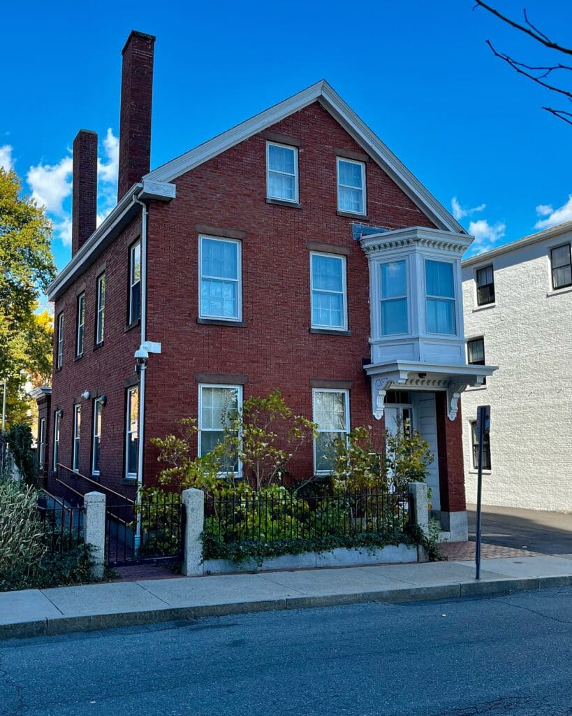 A historic red brick house in Salem, Massachusetts, featuring a white bay window, three chimneys, and a small front garden enclosed by a black iron fence.
