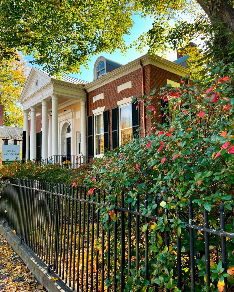 The Salem Athenaeum in Salem, Massachusetts, a historic brick building with white columns, black shutters, and an iron fence surrounded by fall leaves.