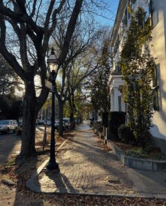 A peaceful brick sidewalk lined with trees and a lamppost in Salem, Massachusetts, with shadows stretching across the path in the late afternoon sunlight. A white building with columns and shutters stands to the right, adding to the charm of the neighborhood.