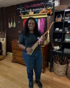 A woman stands inside The Witchery in Salem, Massachusetts, holding a handmade broom decorated with pink twine. Behind her, colorful broom-making materials, baskets, and mystical items are displayed.