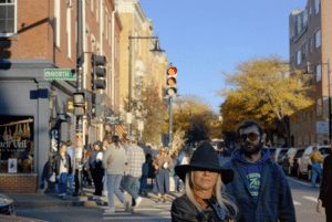 A group of people crossing a street in Salem, Massachusetts at a traffic light, with a woman wearing a black hat in the foreground. Brick buildings, storefronts, and fall foliage can be seen in the background.