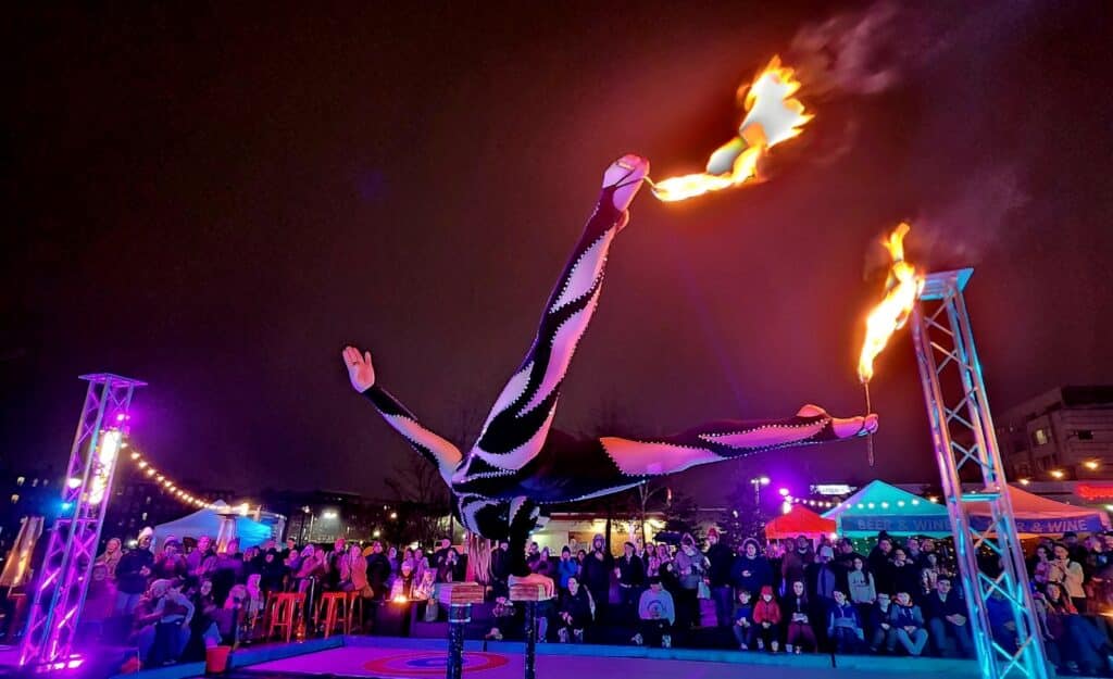 An acrobat performs an incredible handstand on elevated bars while holding flaming props, captivating the audience at a colorful outdoor event in Salem, Massachusetts.