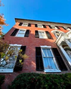A detailed view of the Andrew-Safford House in Salem Massachusetts, featuring red brick walls, tall windows framed with black shutters, and vibrant greenery in the foreground.