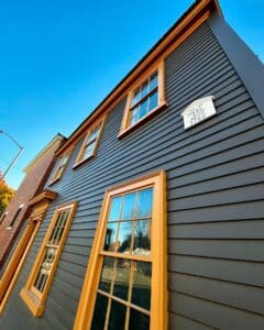 Angled view of the Daniel Bray House in Salem, Massachusetts, displaying its dark siding, golden window trims, and the plaque indicating "Built by Daniel Bray Mariner 1766."