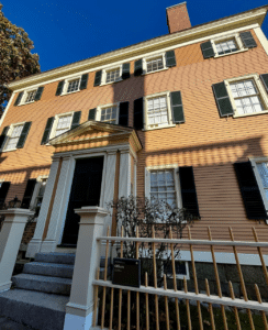 A close-up view of the Hawkes House in Salem, Massachusetts, featuring peach clapboard siding, black shutters, a grand entrance with white columns, and a picket fence under a bright blue sky.
