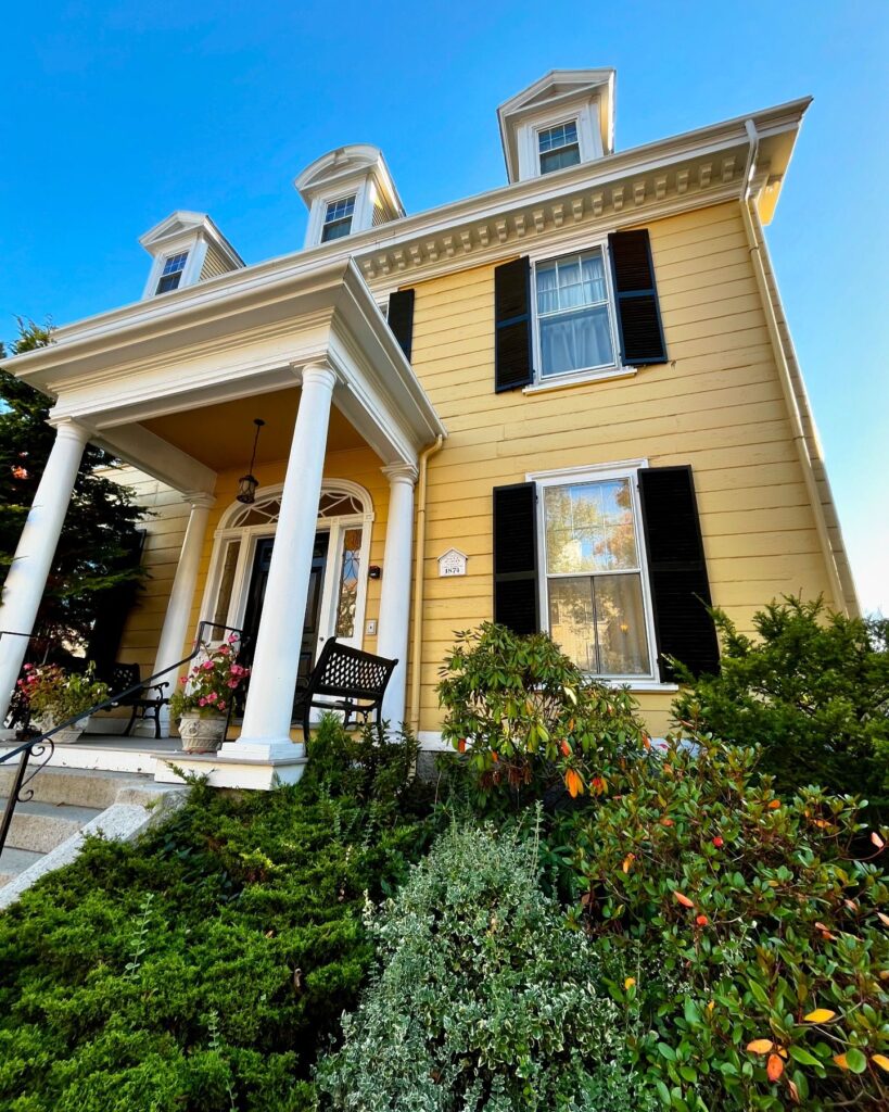 A close-up view of the John P. Peabody House in Salem Massachusetts, showcasing its yellow exterior, white columns, black shutters, and lush greenery around the entrance.