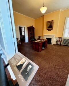 The Collector's Office inside the Salem Custom House in Salem, Massachusetts, featuring period furnishings, a historic desk, a fireplace, and portraits displayed on a placard in the foreground.