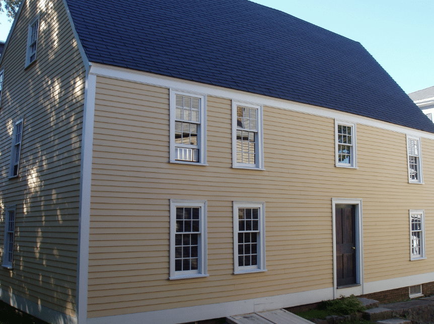 The image shows the Gedney House, a historic First Period home located in Salem, Massachusetts. The house, constructed in 1665 by shipbuilder Eleazer Gedney, features traditional colonial architecture with its steeply pitched roof and wooden clapboard exterior. The yellow-painted structure is complemented by a white picket fence, adding to its charm. In the background, a smaller red building with a central chimney is visible, reflecting the architectural styles of the era. The Gedney House is now managed by Historic New England and is preserved as a testament to Salem's colonial history and evolving architectural practices.