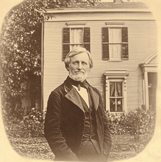 A sepia-toned photograph of Dr. Nathaniel Peabody standing in front of a traditional 19th-century New England home. He is dressed formally in a suit and bow tie, exuding an air of dignity and sophistication. The house behind him features wooden siding, shuttered windows, and a classic architectural style, reflecting the period. The image captures Dr. Peabody's presence as a respected figure in his community and offers a glimpse into the environment in which he lived and worked.