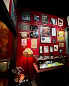 A corner of the Salem Witch Board Museum in Salem, Massachusetts, featuring a red wall adorned with framed Ouija board memorabilia, vintage advertisements, and photographs. Below the wall is an antique display case holding Ouija boards and related artifacts, alongside a vintage lamp and decorative plant.