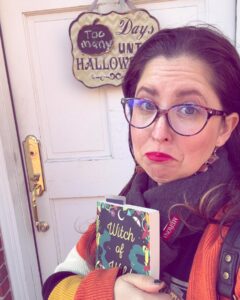 A person in Salem, Massachusetts, wearing glasses and a cozy scarf stands in front of a sign that reads "Too Many Days Until Halloween" while holding a book titled "Witch of Wildwood."