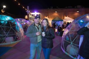 A smiling couple holds drinks while standing among illuminated igloos and festive tents at the Salem Frozen Fire Festival in Salem, Massachusetts.