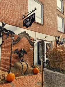 The storefront of Coven - A Dark Boutique, featuring Halloween decorations with skeleton figures, pumpkins, and an archway under a black store sign that reads "COVEN" in Salem Massachusetts